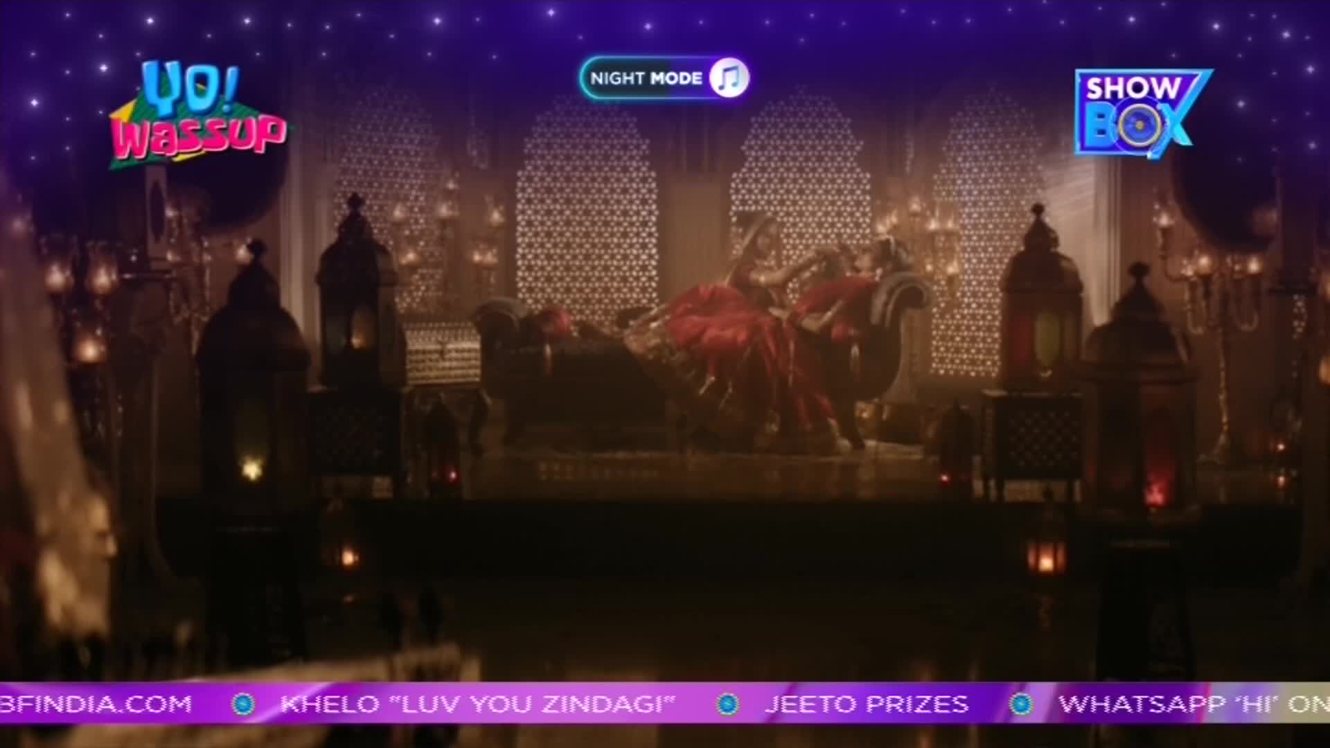 On a stage lit by lanterns, a couple in ornate red clothing reclines on a dark chaise lounge. The scene appears to be part of a production, likely on ShowBox, with the "Yo! Wassup" logo visible.
