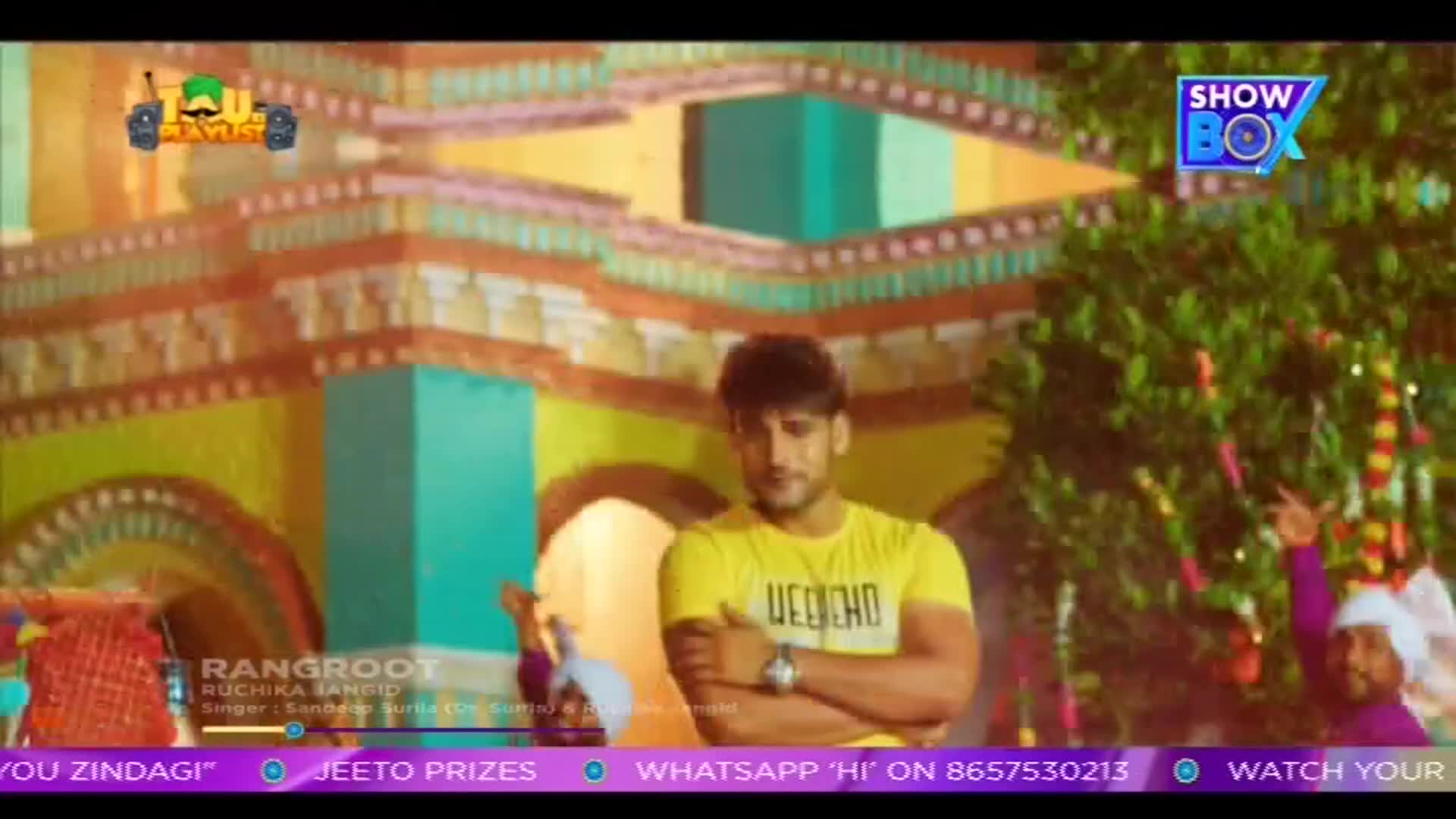 A man in a yellow t-shirt stands with crossed arms, looking down, while a brightly decorated structure looms behind him. The ShowBox logo is visible, and the screen displays a music video from India.
