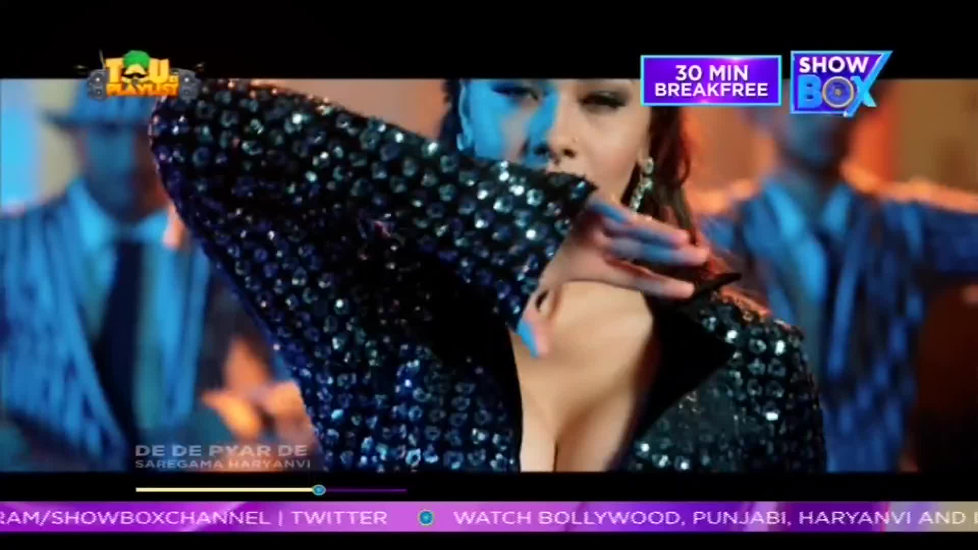 A woman in a sequined jacket is reaching out, her arm bent across her face. Behind her, a few men in suits are blurred, all on a ShowBox music video from India.
