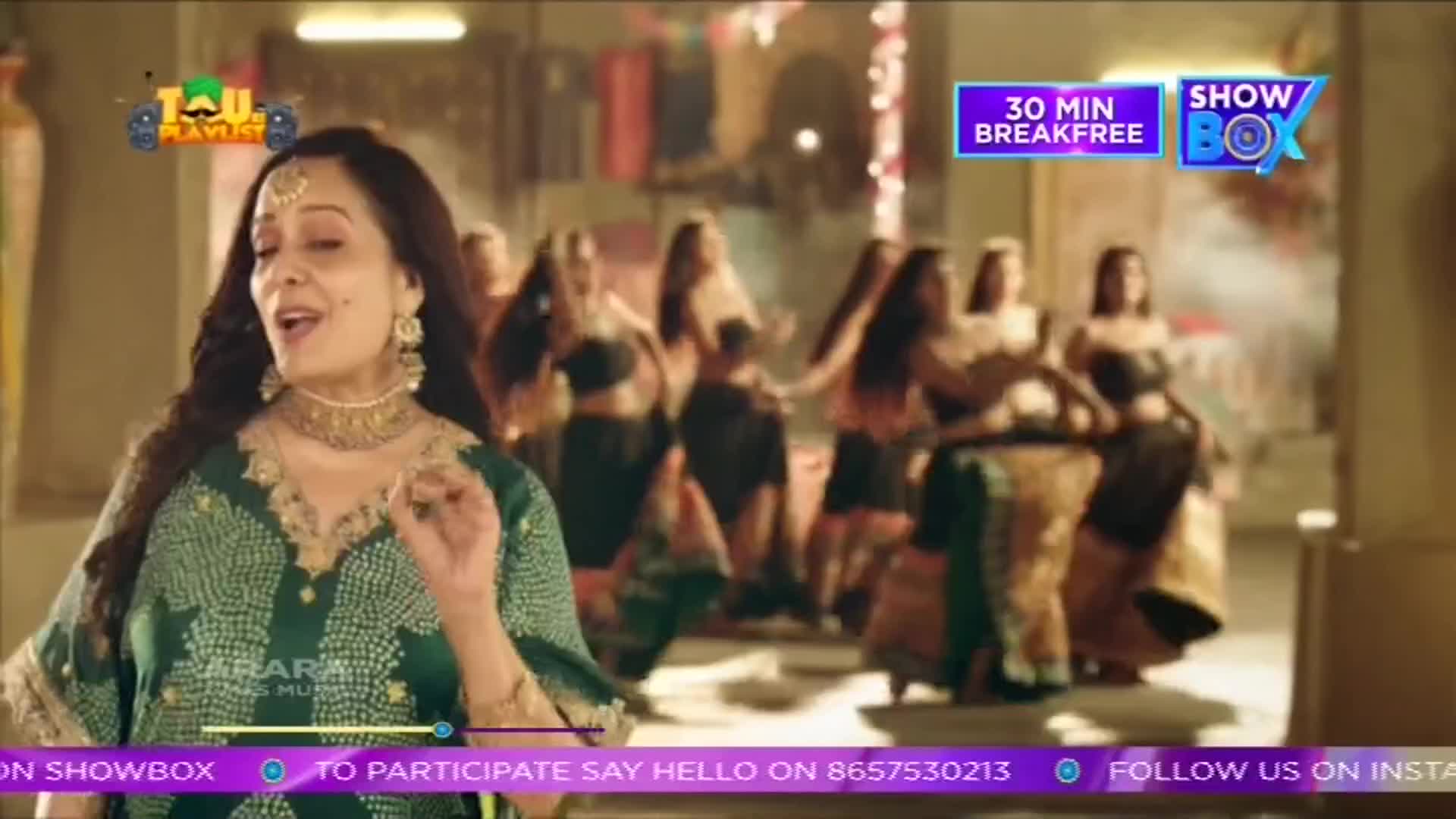 A woman in a green dress sings, gesturing with her hand, while a group of dancers sway in the background. The scene looks like a music video, possibly from India, with a ShowBox logo in the corner.
