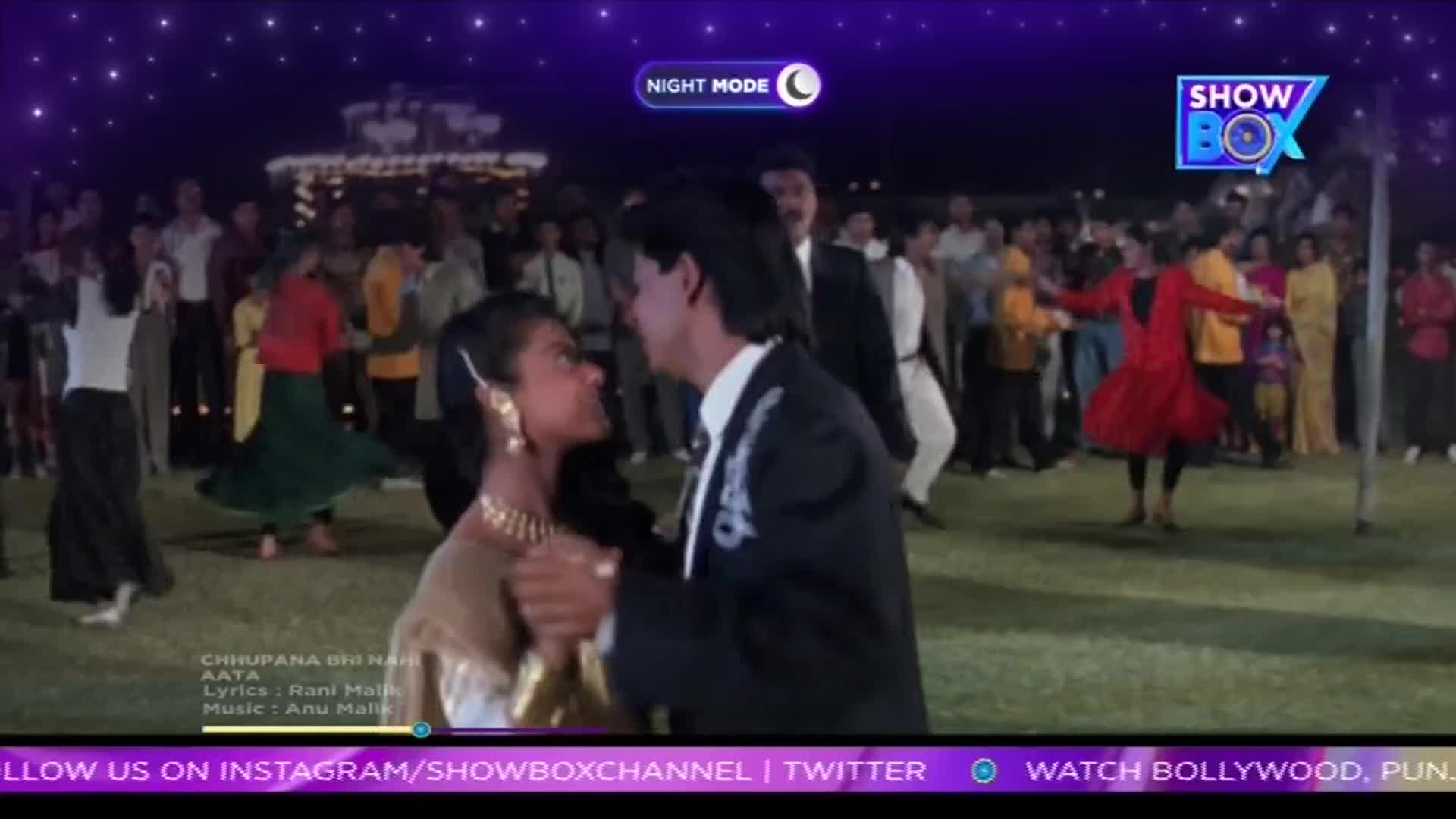 A couple dances close, while others swirl and twirl on the grass, illuminated by the night mode of ShowBox. The scene is from a Bollywood film, with the song lyrics displayed at the bottom of the screen.
