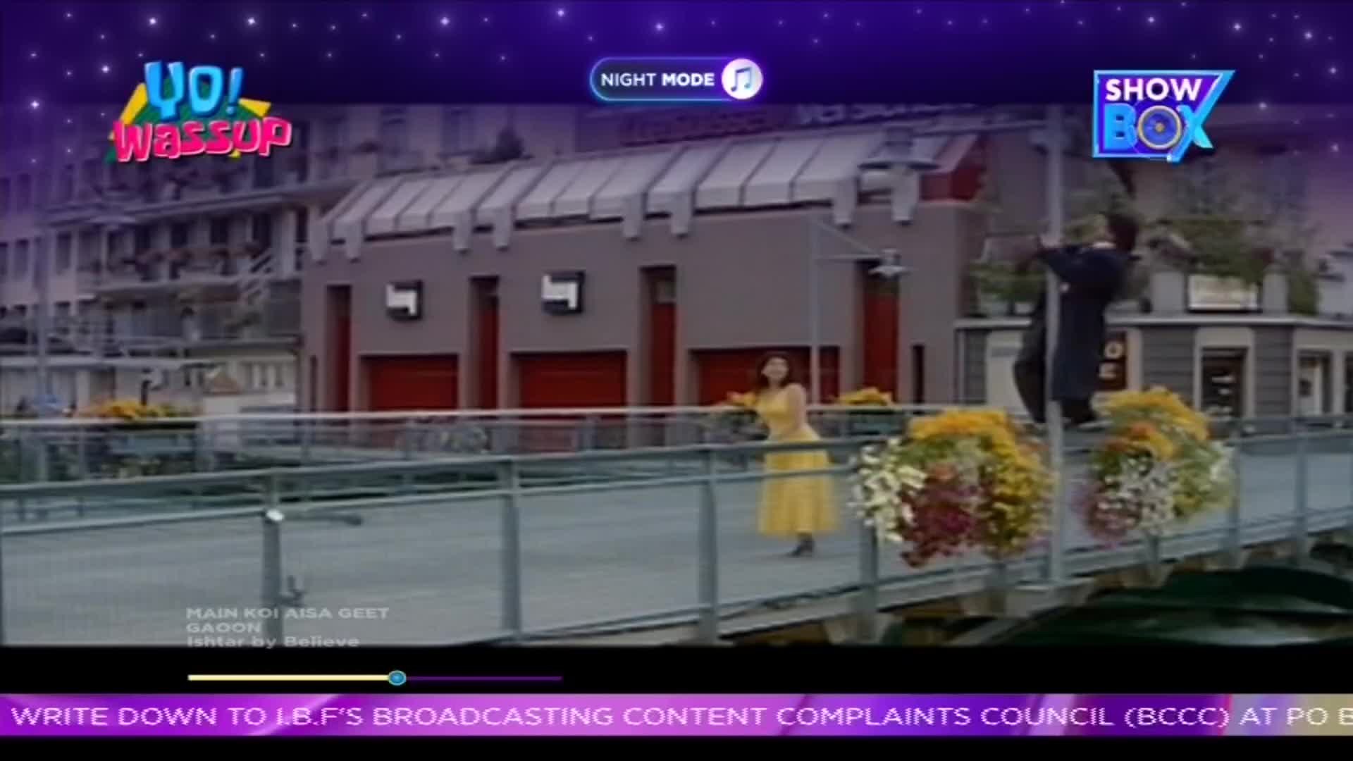 A woman in a yellow dress is dancing on a bridge, while a man in a dark suit gestures dramatically nearby. The scene is playing on ShowBox, with a "Yo! Wassup" logo in the corner.
