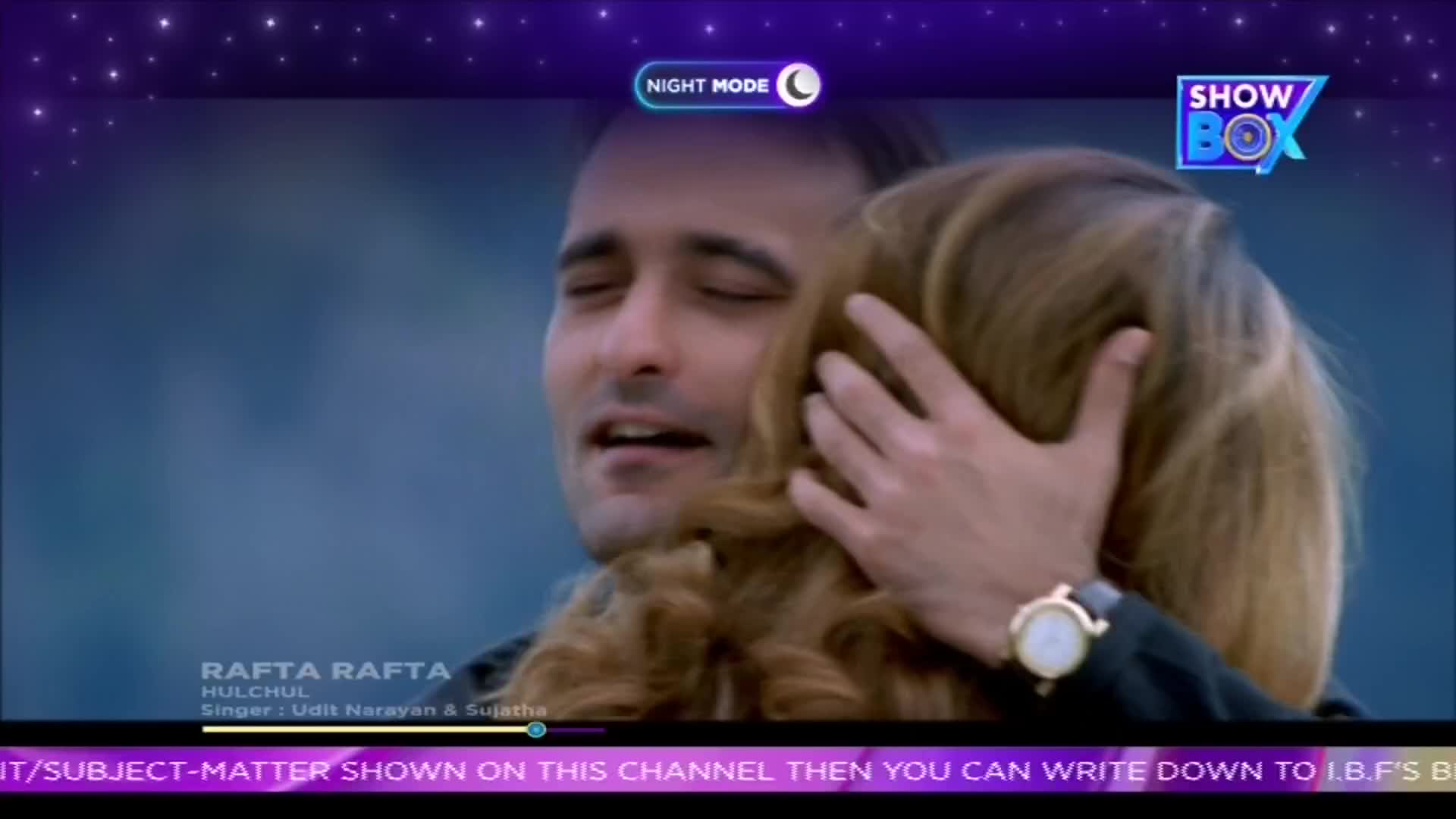 A man, eyes closed, embraces a woman, her blonde hair cascading over his shoulder. The scene appears on ShowBox, a channel from India, with song lyrics displayed at the bottom.
