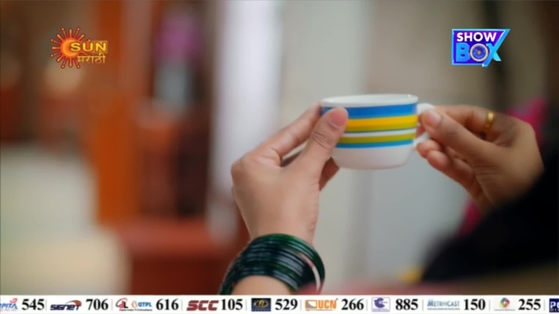 A woman's hands cradle a small, striped cup, the handle held between her fingers.  Green bangles adorn her wrist, and a television logo for ShowBox is visible in the background.
