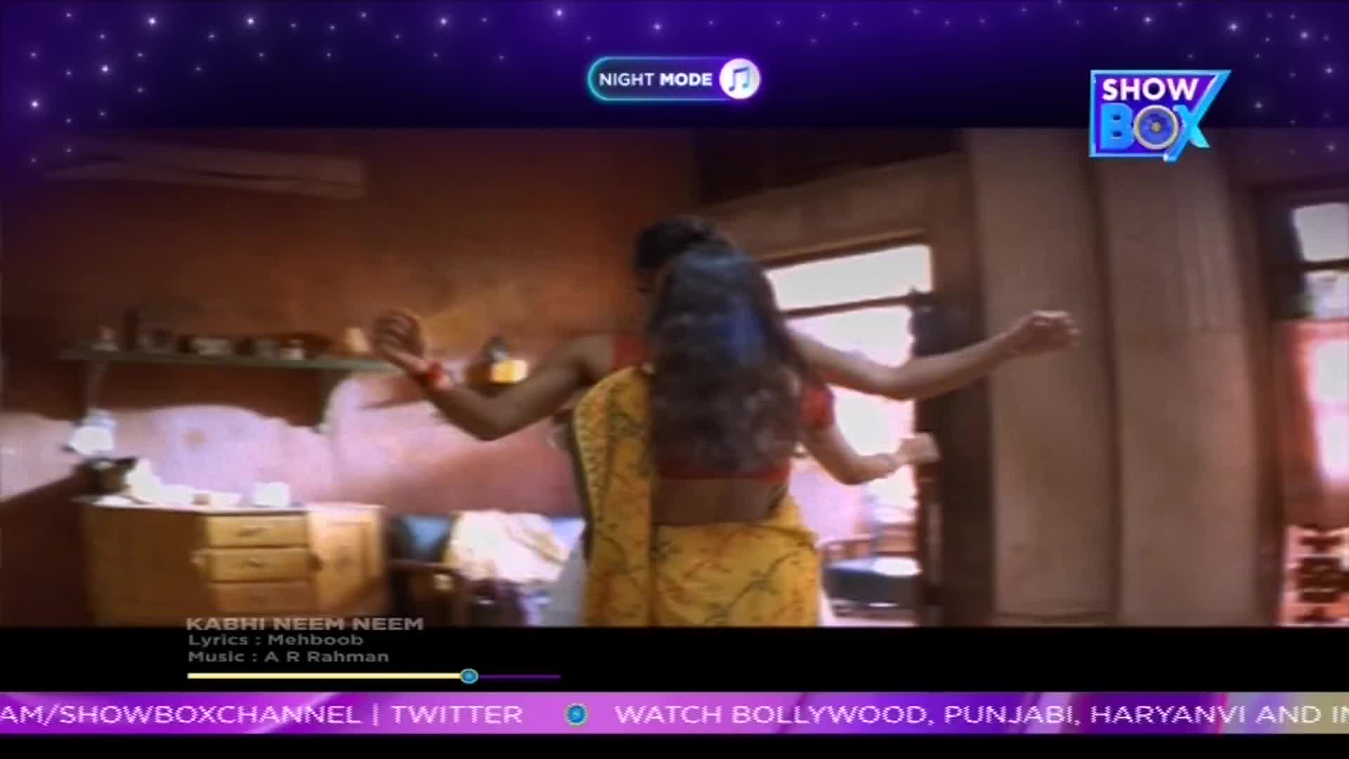 A woman in a yellow sari dances with her arms outstretched, her back to the camera. The scene is from a music video on ShowBox, with the lyrics and music credits displayed at the bottom.
