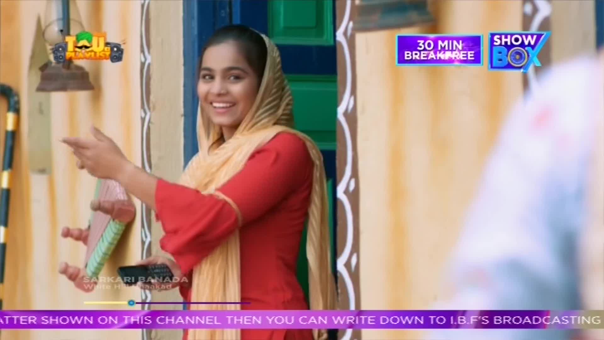 A woman in a red outfit and headscarf gestures enthusiastically, holding a remote control. The ShowBox logo is visible in the background, along with a logo for "Sarkari Banada."
