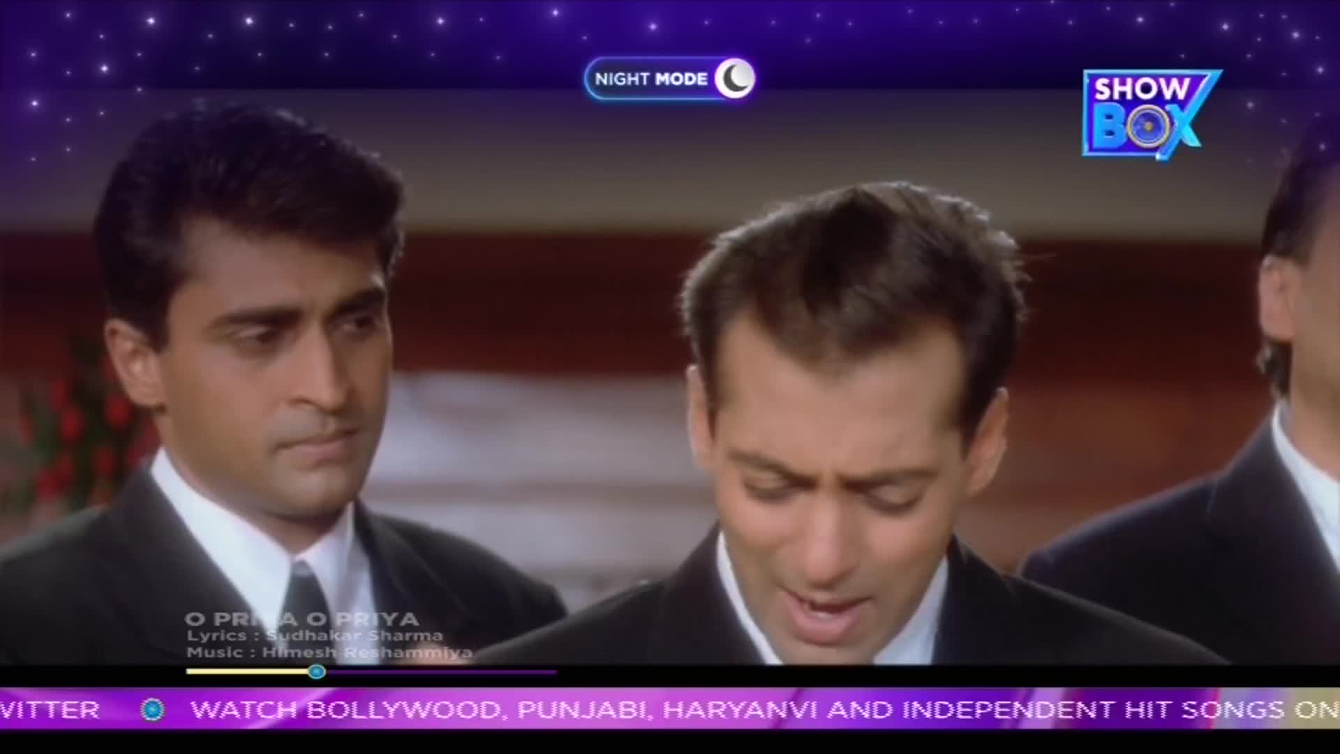 Two men in suits are standing close together, with one singing and the other looking on with a slight frown. The ShowBox logo is in the corner, and the song's title and credits are displayed on the screen.
