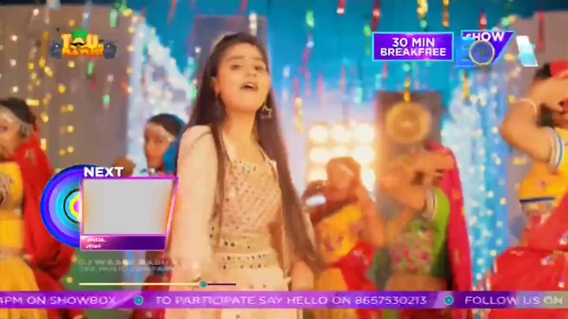 A young woman in a sequined dress sings, arms raised, surrounded by dancers in colorful costumes. The scene is from a ShowBox production, with graphics and text overlaying the performance.
