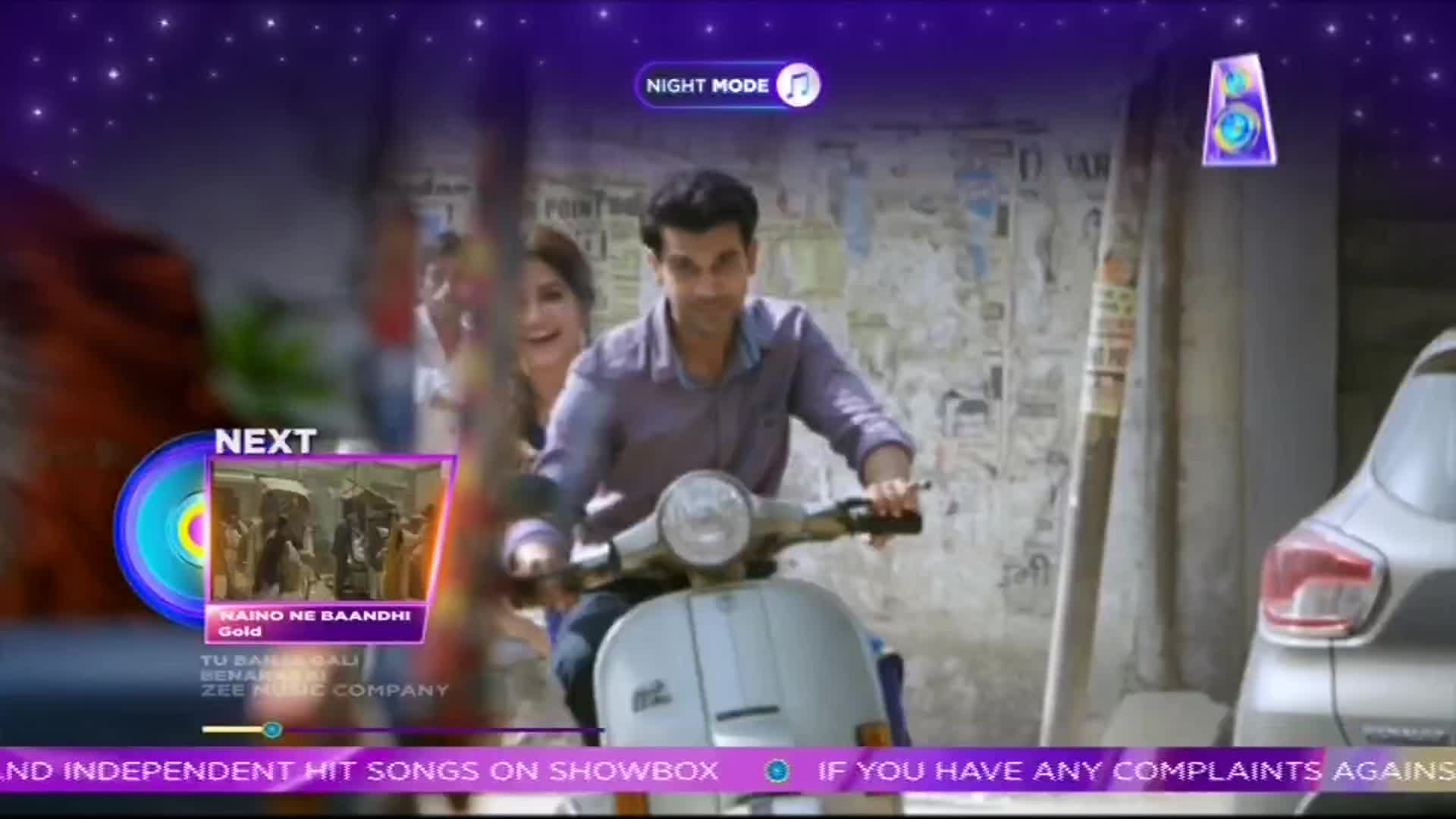 A man steers a light blue scooter down a street, with a woman smiling behind him. The ShowBox logo and a "Next" graphic with a song title overlay the scene, as if it's a music video.
