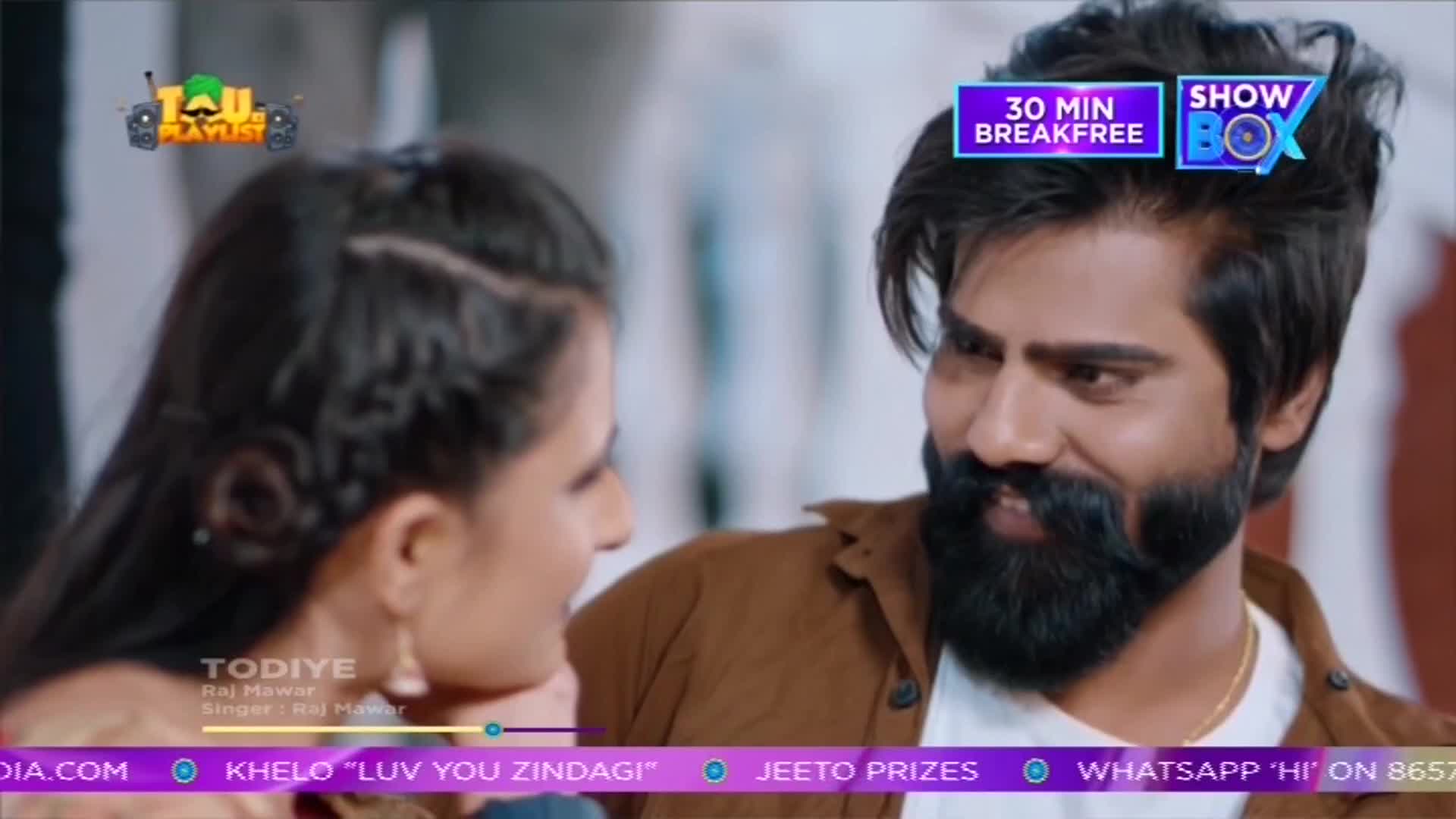 A man with a thick beard and a woman are gazing at each other, their faces close. The scene seems to be part of a music video, with text from ShowBox and India visible at the top and bottom of the screen.
