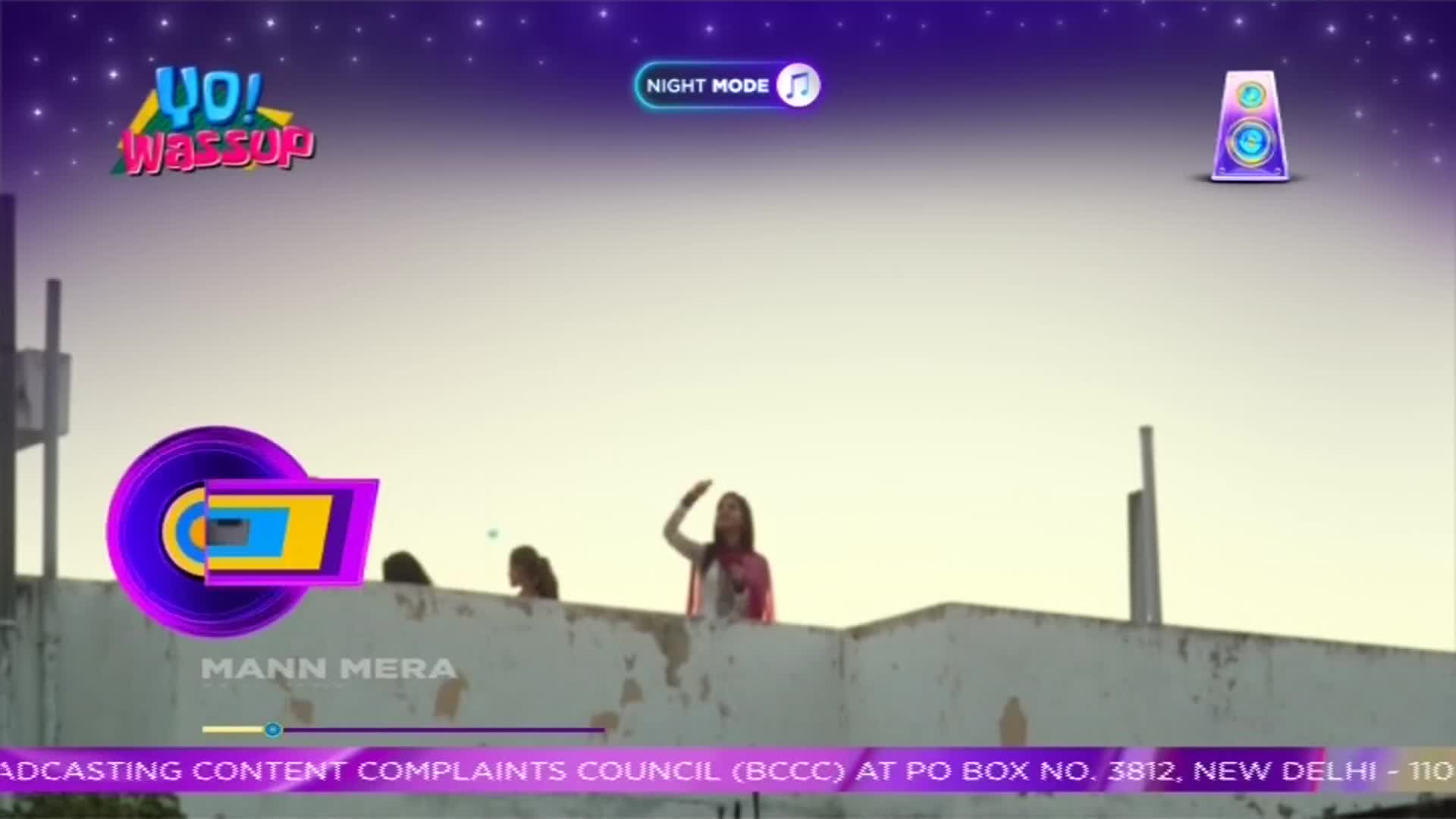 On a rooftop, a woman in a pink scarf waves her hand towards the sky. The screen displays the "Yo! Wassup" logo, a music video from India, and the words "Mann Mera."
