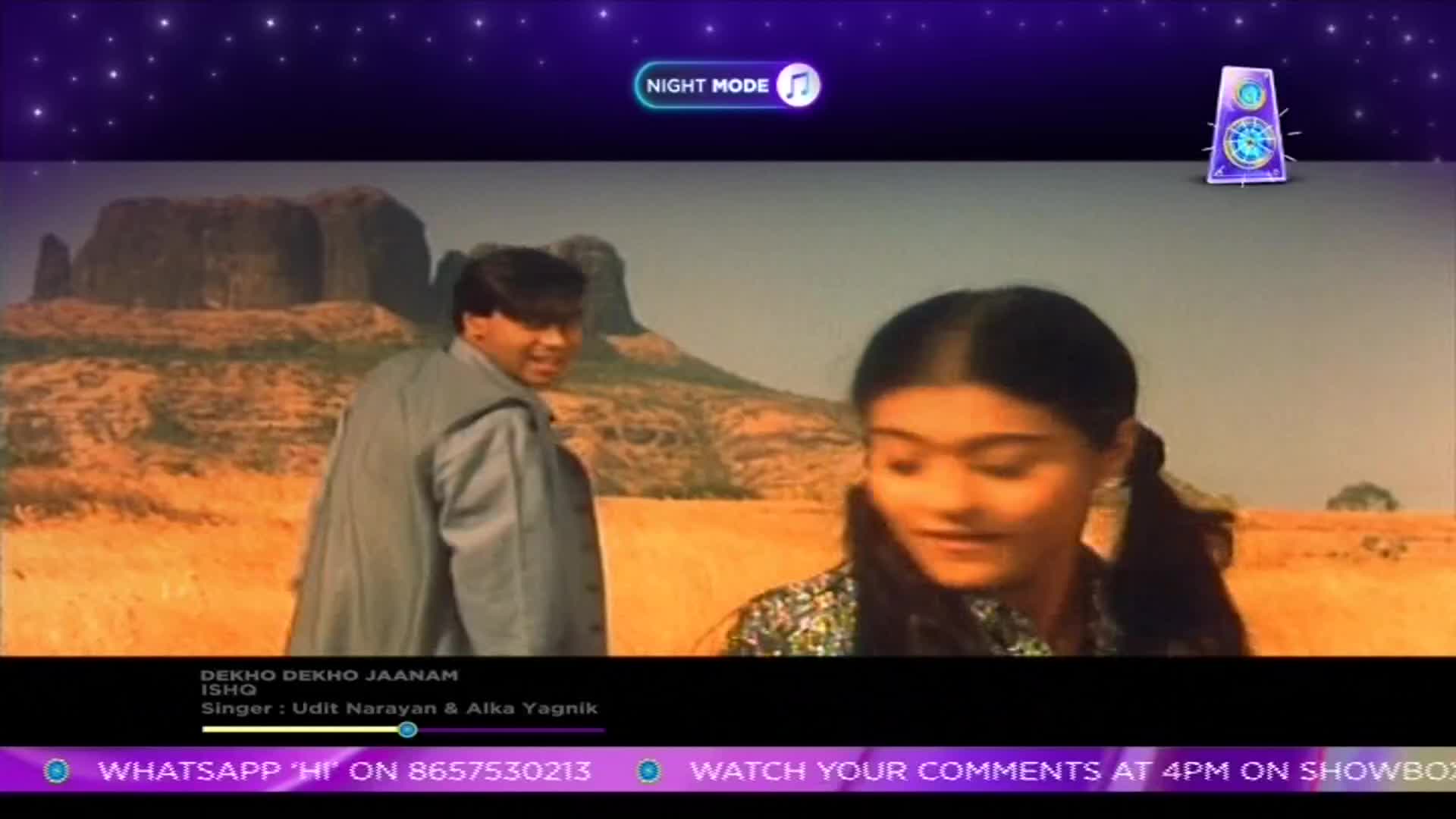 A man in a grey jacket turns his head to look at a woman with pigtails in a dry, grassy landscape. The song "Dekho Dekho Jaanam" plays, a familiar tune from Indian cinema.