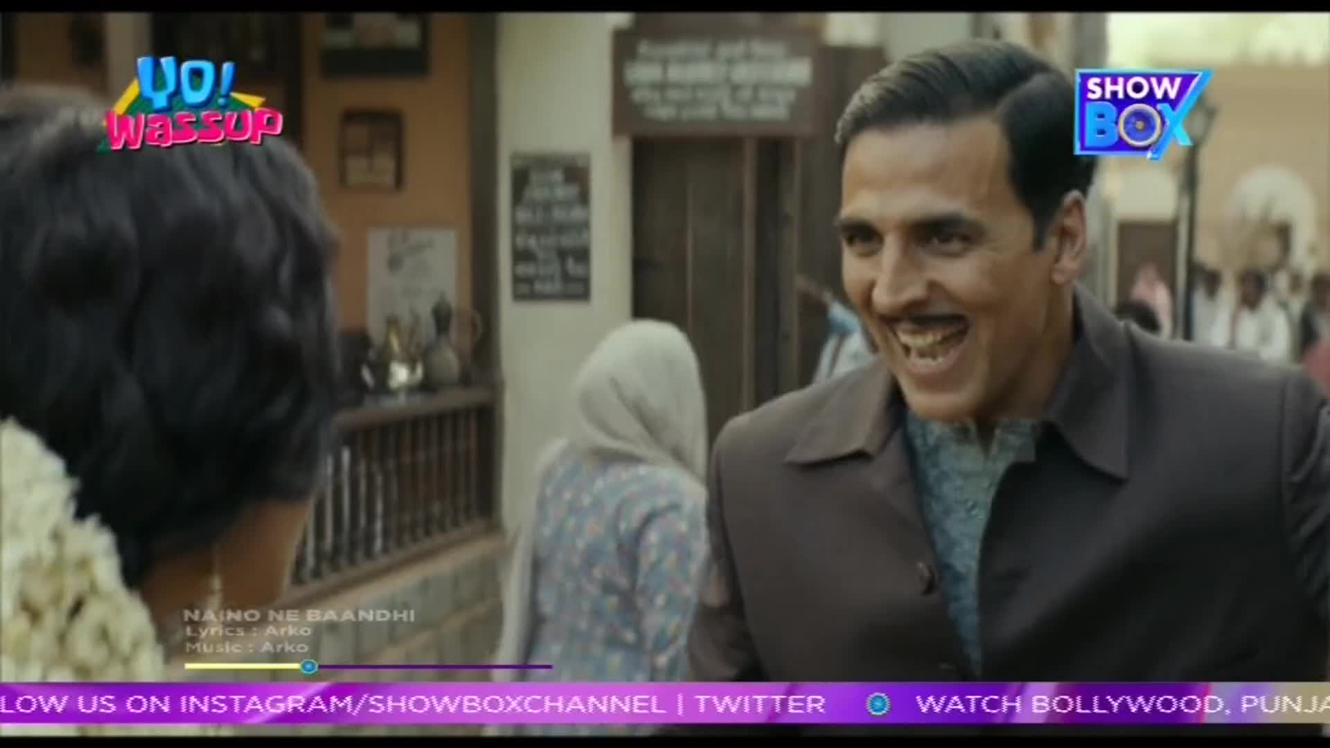 A man with a wide, toothy grin laughs heartily, his eyes crinkling at the corners. Behind him, a woman in a headscarf walks away, and the bustling atmosphere of an Indian street unfolds.