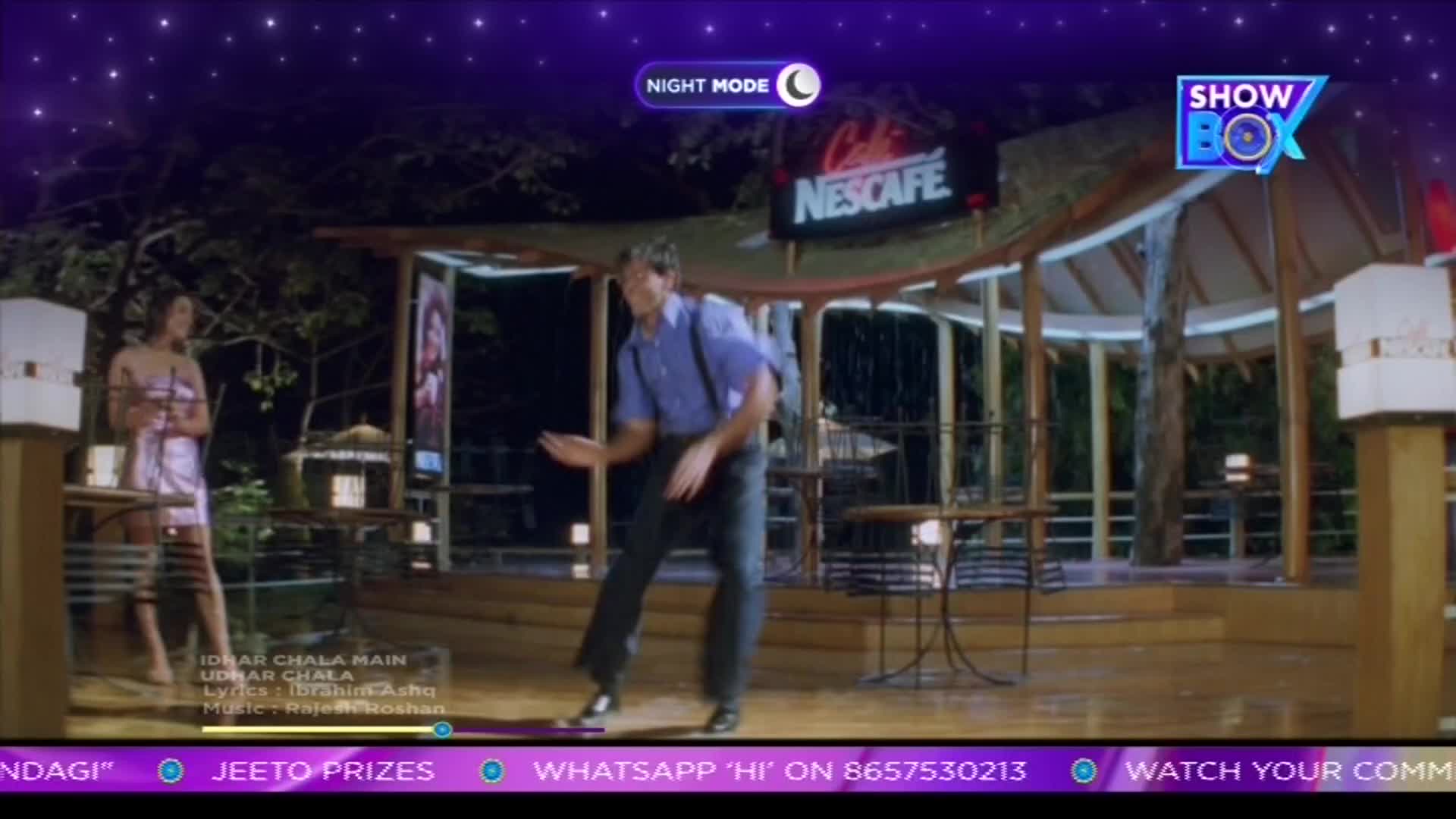 A man in a blue shirt and dark pants dances energetically under a covered outdoor cafe. A woman in a shimmering dress watches him from a short distance.