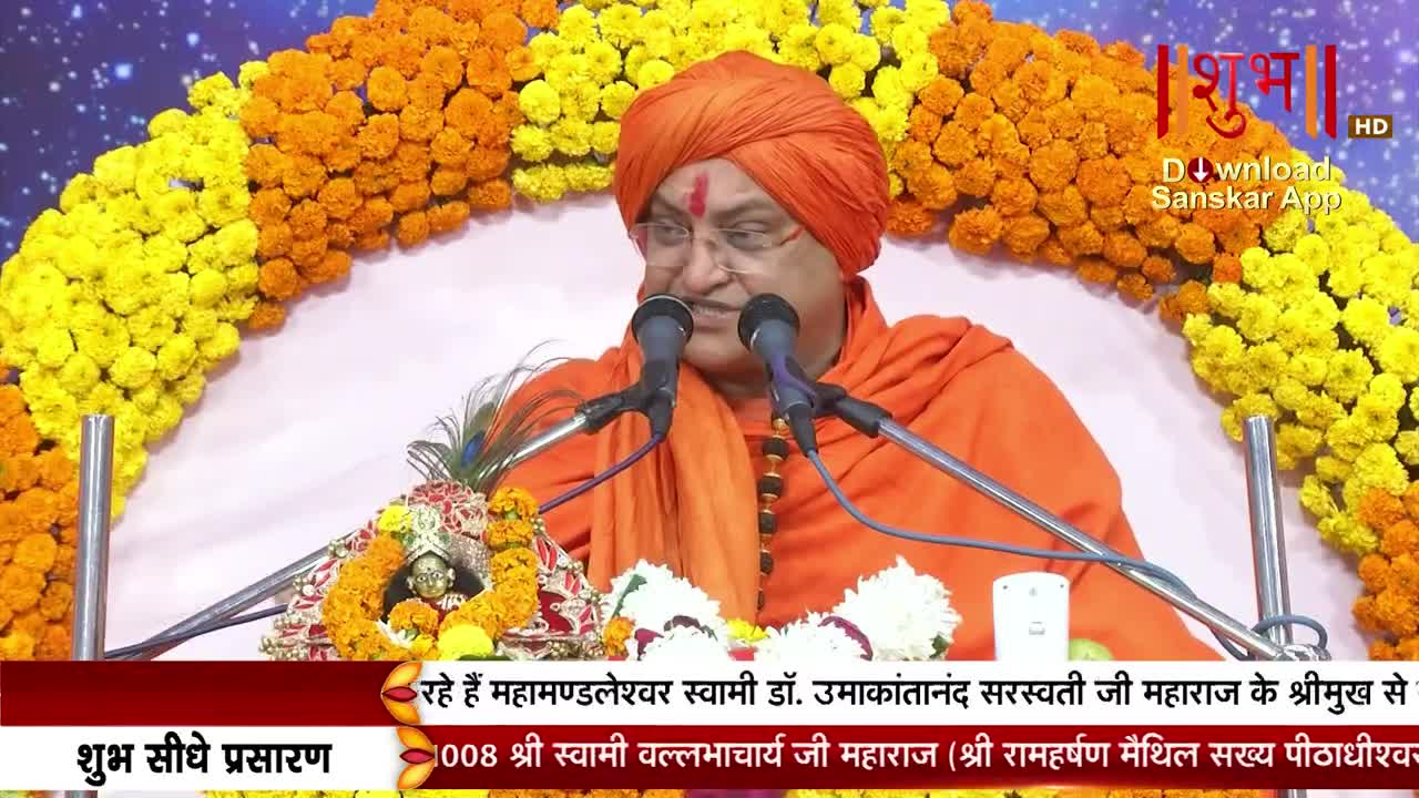A man in an orange turban and robes speaks into two microphones, his face framed by bright yellow and orange flowers. Below, text scrolls across the screen, identifying the speaker and the broadcast on Shubh TV.
