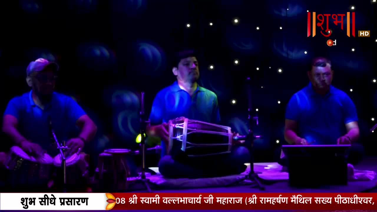 Three men in blue shirts are seated, playing instruments on a stage. The central musician is striking a large drum, while the others keep time on smaller drums and a keyboard, all under the glow of stage lights, as Shubh TV broadcasts the performance.
