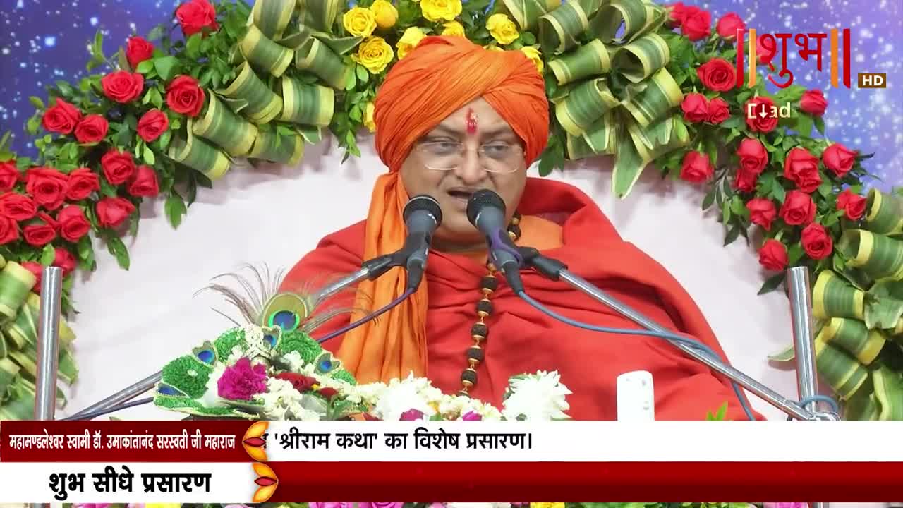 A robed man with an orange turban speaks into two microphones, a beaded necklace resting on his chest. The scene is framed by vibrant flower arrangements, and the Shubh TV logo is visible in the top right.
