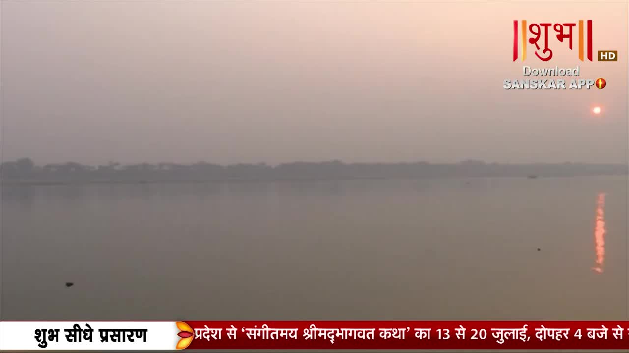 The sun, a hazy orange disc, hangs low over the water, its reflection shimmering across the calm surface. A broadcast from Shubh TV is showing a wide shot of the river, with a distant treeline barely visible through the morning mist.
