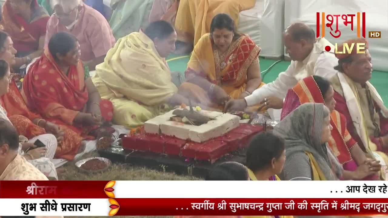 People in colorful clothing sit on the floor around a small, square fire pit, some reaching toward it. The scene is being broadcast live on Shubh TV, with text scrolling across the bottom of the screen.
