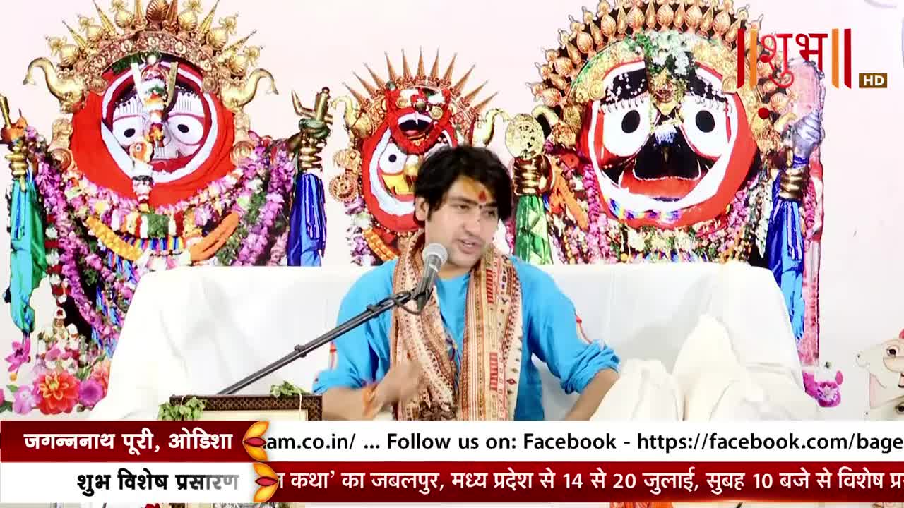 A man in a blue shirt speaks into a microphone, gesturing with his hands. Behind him, two ornate, colorful religious icons are displayed on a white platform, as seen on Shubh TV.
