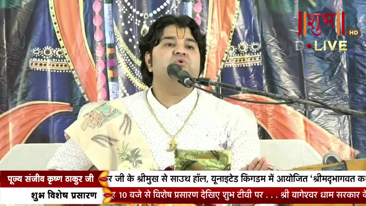 A man in a white shirt speaks into a microphone, his words broadcast live on Shubh TV. Behind him, a colorful backdrop hangs, and text scrolls across the bottom of the screen.
