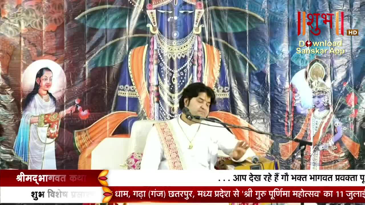 A man in white robes speaks into a microphone as he sits on a stage. Behind him, a backdrop features religious figures, and the Shubh TV logo is visible in the corner.
