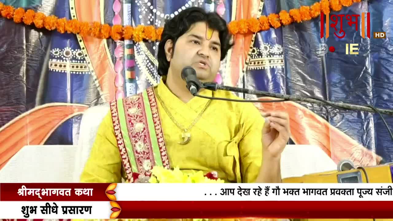 A man in a yellow shirt speaks into a microphone, his hand gesturing as he addresses the audience. The scene is being broadcast live on Shubh TV, with text in Hindi at the bottom of the screen.
