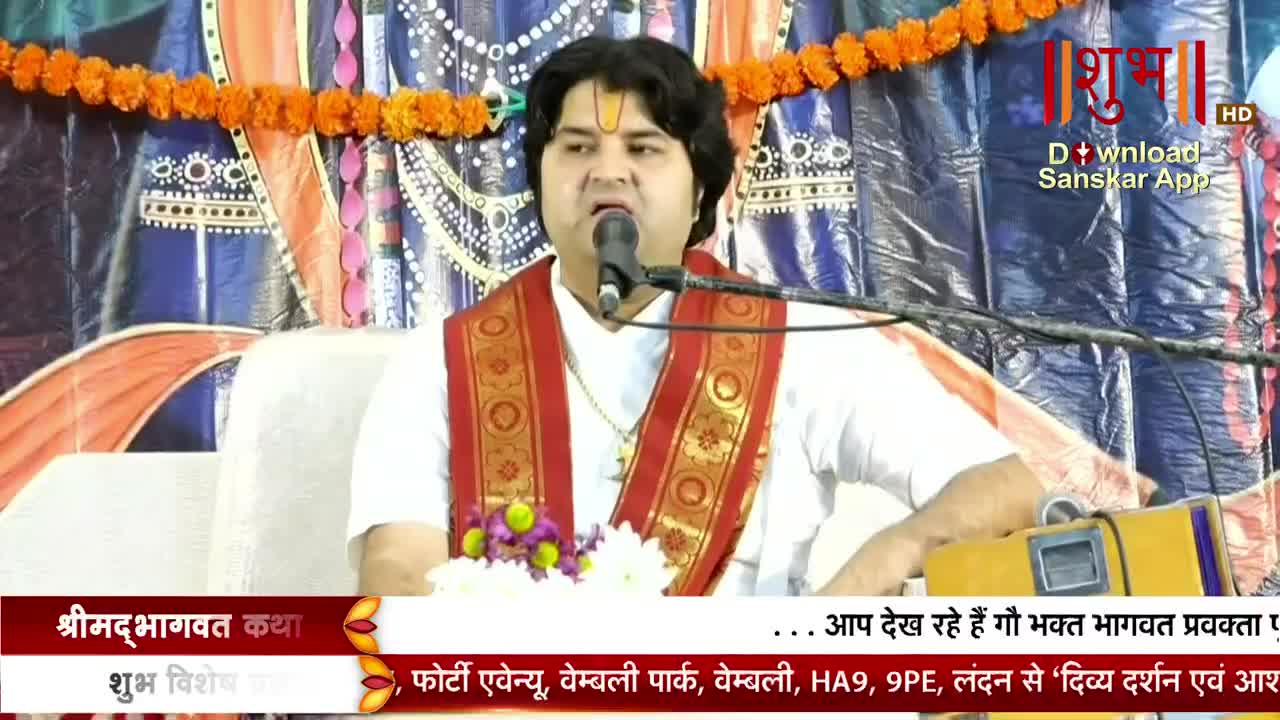 A man in a white kurta speaks into a microphone, his face lit by the stage lights. He's wearing a red and gold scarf, and the Shubh TV logo is visible in the corner.
