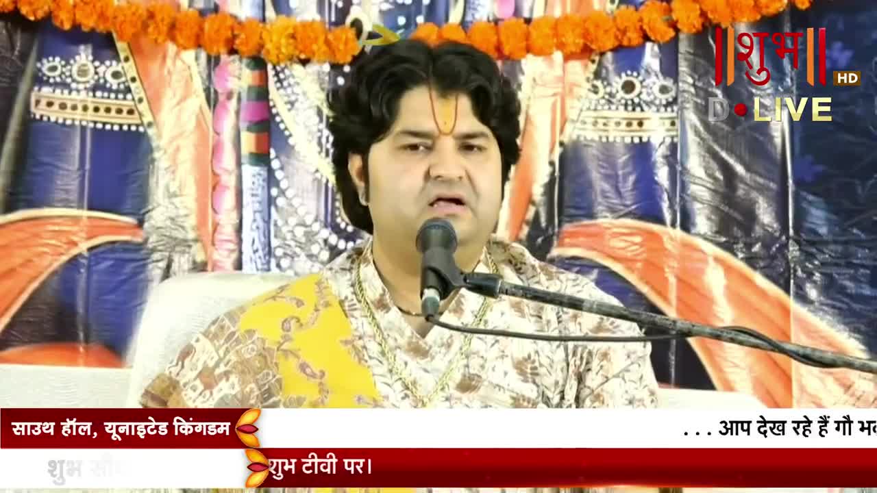 A man in a patterned shirt speaks into a microphone, his face illuminated by the stage lights. The broadcast is live on Shubh TV, with a banner reading "South Hall, United Kingdom" at the bottom of the screen.

