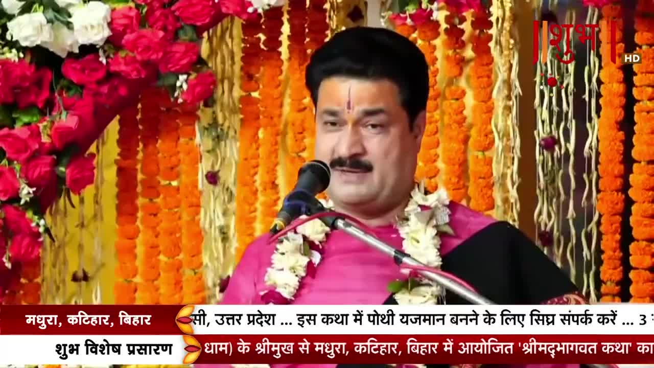 A man with a prominent mustache speaks into a microphone, a garland draped around his neck. Behind him, marigold garlands and floral arrangements decorate the stage, and the Shubh TV logo is visible in the corner.
