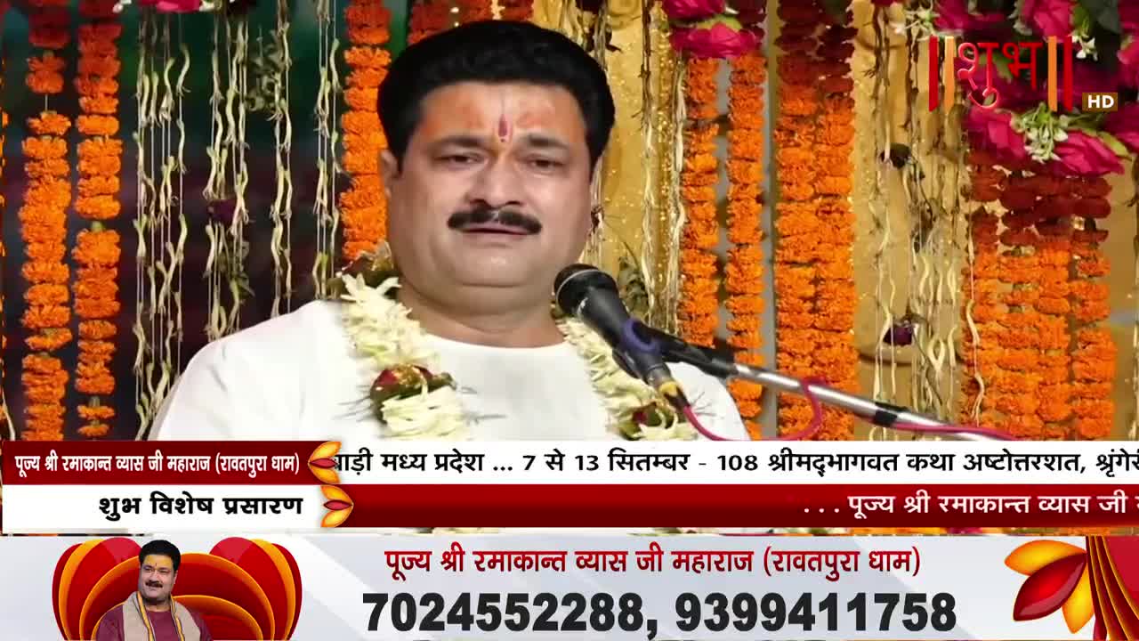 A man in a white shirt speaks into a microphone, a garland around his neck, likely for a Shubh TV broadcast. Behind him, strings of orange flowers and garlands hang, creating a warm, festive backdrop.
