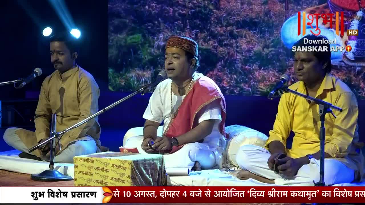 Three men sit cross-legged on a stage, each with a microphone. The central figure, wearing a red scarf, sings into his mic, while the others listen intently.
