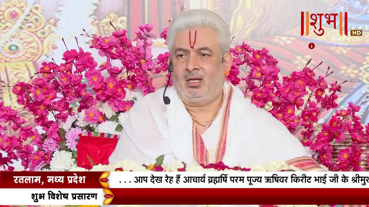 A man with white hair and a red mark on his forehead speaks into a microphone, surrounded by bright pink flowers. The Shubh TV logo is visible, and text at the bottom indicates a broadcast from Ratlam, Madhya Pradesh, India.
