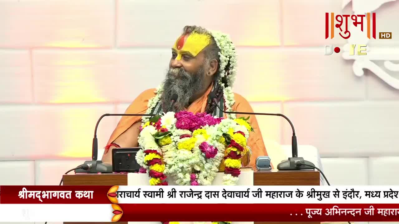 A man with a long beard and a garland of flowers sits behind a lectern, speaking. The Shubh TV logo is visible in the corner, and a banner with Hindi text runs along the bottom of the screen.

