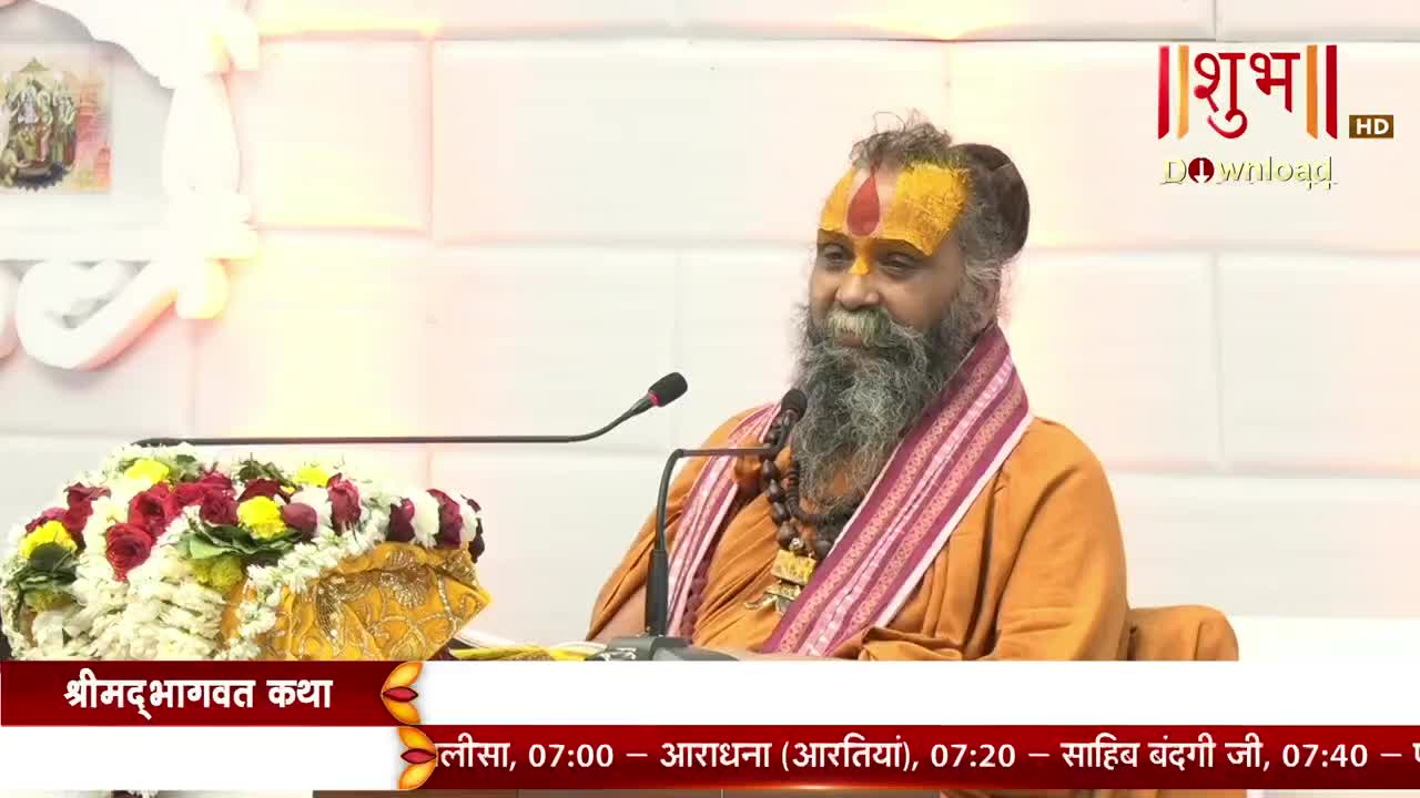 A man with a long, gray beard and saffron markings sits at a desk, speaking into a microphone. The Shubh TV logo is visible in the top right corner, and the screen displays text about a program called "Shrimad Bhagwat Katha."
