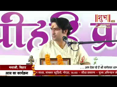 A man in a cream-colored shirt speaks into a microphone on a Shubh TV broadcast from India. Behind him, large purple Hindi text is visible, along with a small framed image and floral decorations.
