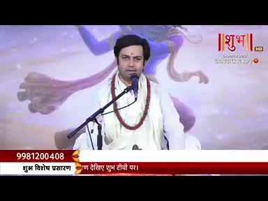 A man in white robes speaks into a microphone, a red bead necklace resting on his chest. The Shubh TV logo is visible on the screen behind him, and a phone number is displayed below.
