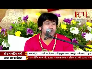 A man in a red shirt speaks into a microphone, framed by flowers and the logo of Shubh TV. The lower third of the screen displays text in Hindi, listing program details.
