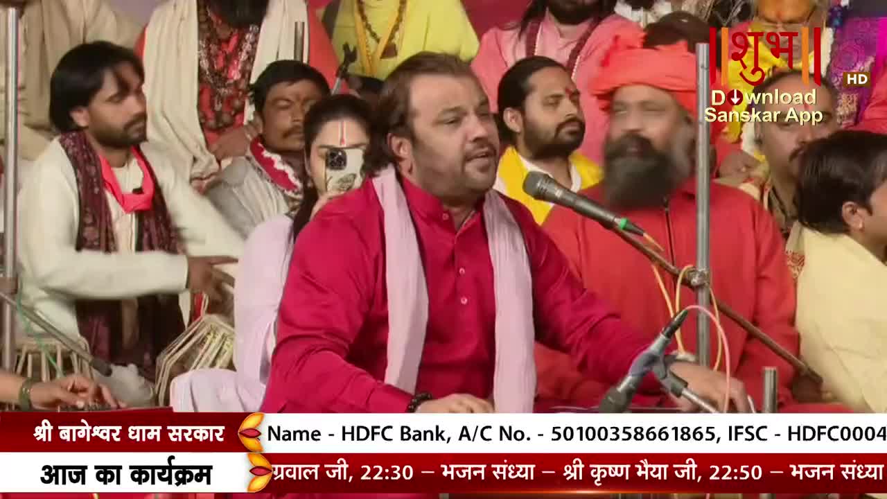 A man in a red kurta, draped with a white scarf, speaks into a microphone, his expression animated. Behind him, a group of people, some in religious attire, listen intently, and the Shubh TV logo is visible in the corner.
