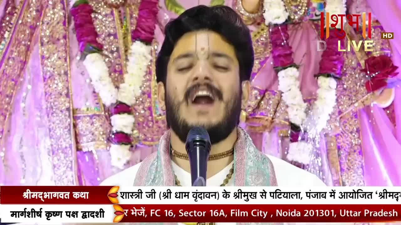 A man with a small, dark beard sings into a microphone, his eyes closed. Behind him, draped fabric and garlands of flowers create a backdrop, and the Shubh TV logo is visible in the corner.

