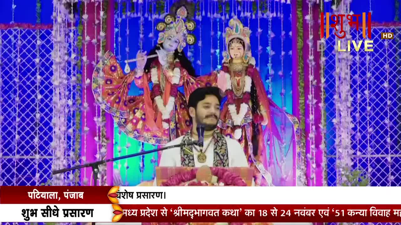 A man in a white shirt stands at a podium, speaking into a microphone. Behind him, decorated statues of Krishna and Radha are illuminated on a stage, with the Shubh TV logo visible in the corner.
