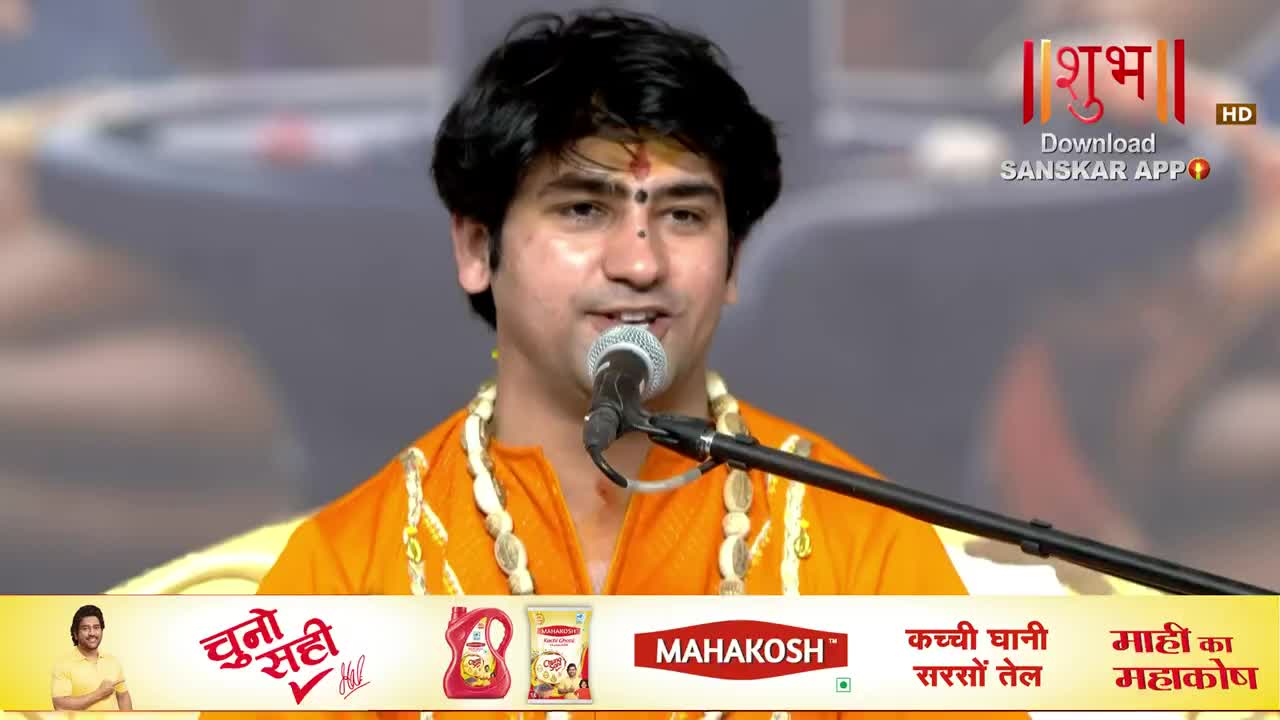 A man in an orange outfit speaks into a microphone, a small red mark on his forehead. The Shubh TV logo is visible in the corner, and a commercial banner runs along the bottom.

