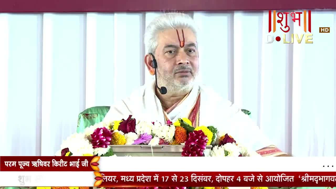 A man in white robes sits before a floral arrangement, speaking into a small microphone. The Shubh TV logo is visible in the upper right corner, and text in Hindi scrolls at the bottom of the screen.
A man in white robes sits before a floral arrangement, speaking into a small microphone. The Shubh TV logo is visible in the upper right corner, and text in Hindi scrolls at the bottom of the screen.