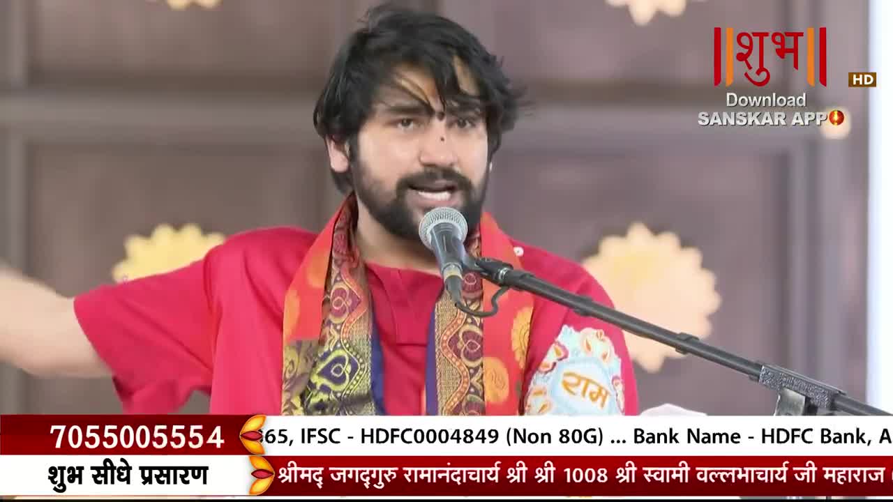 A man in a red shirt and scarf is speaking into a microphone, gesturing with his left hand. The Shubh TV logo and contact information are visible in the corner of the screen.
A man in a red shirt and scarf is speaking into a microphone, gesturing with his left hand. The Shubh TV logo and contact information are visible in the corner of the screen.
