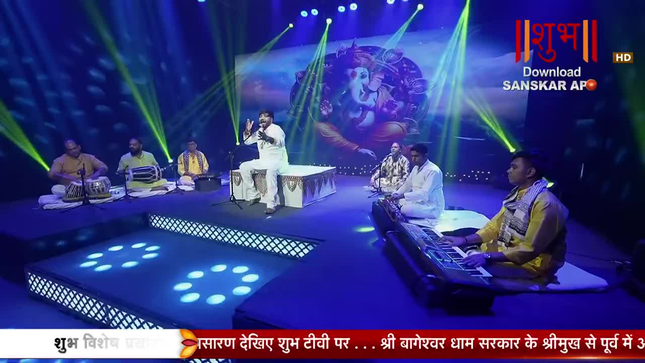 A singer in white sits on a raised platform, gesturing with his hands as a group of musicians accompanies him. One man plays a keyboard, while others tap on tabla drums and a mridangam.
