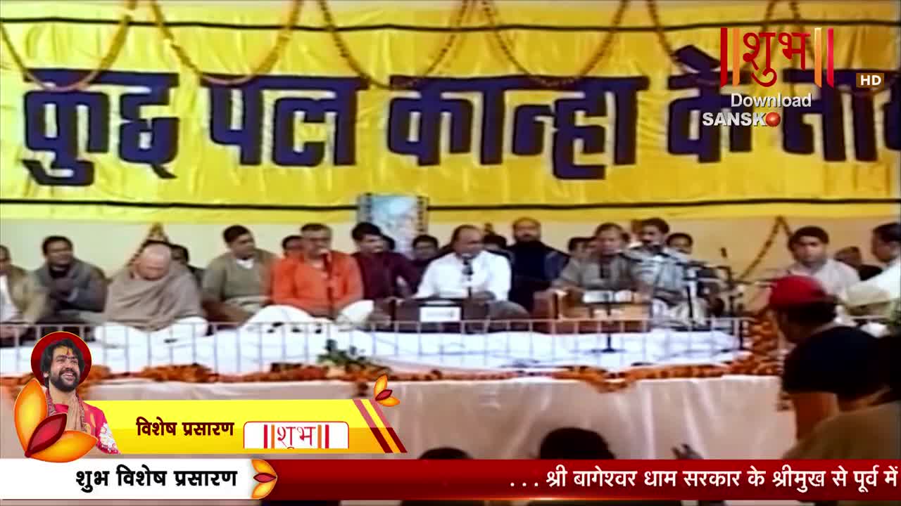 A group of men sit on a raised platform, some singing into microphones. A large yellow banner with Hindi text hangs behind them.
