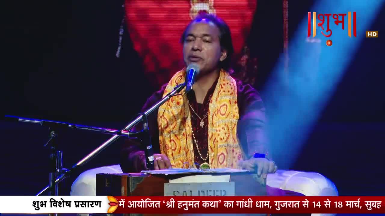 A man in a maroon outfit and a yellow scarf sings into a microphone. He's seated at a harmonium, with a bright blue light shining on him.