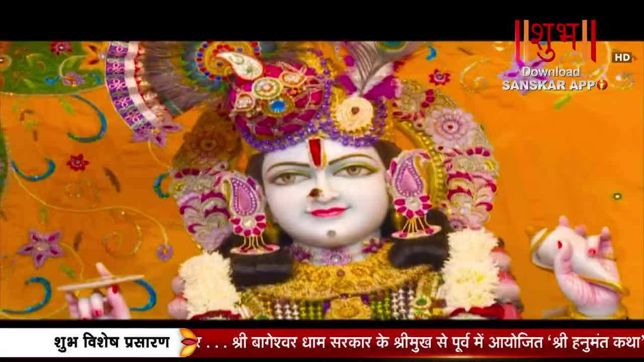 A statue of a deity, adorned with a jeweled crown and floral garlands, is centered in the frame. The backdrop is a vibrant orange fabric with intricate gold embroidery.