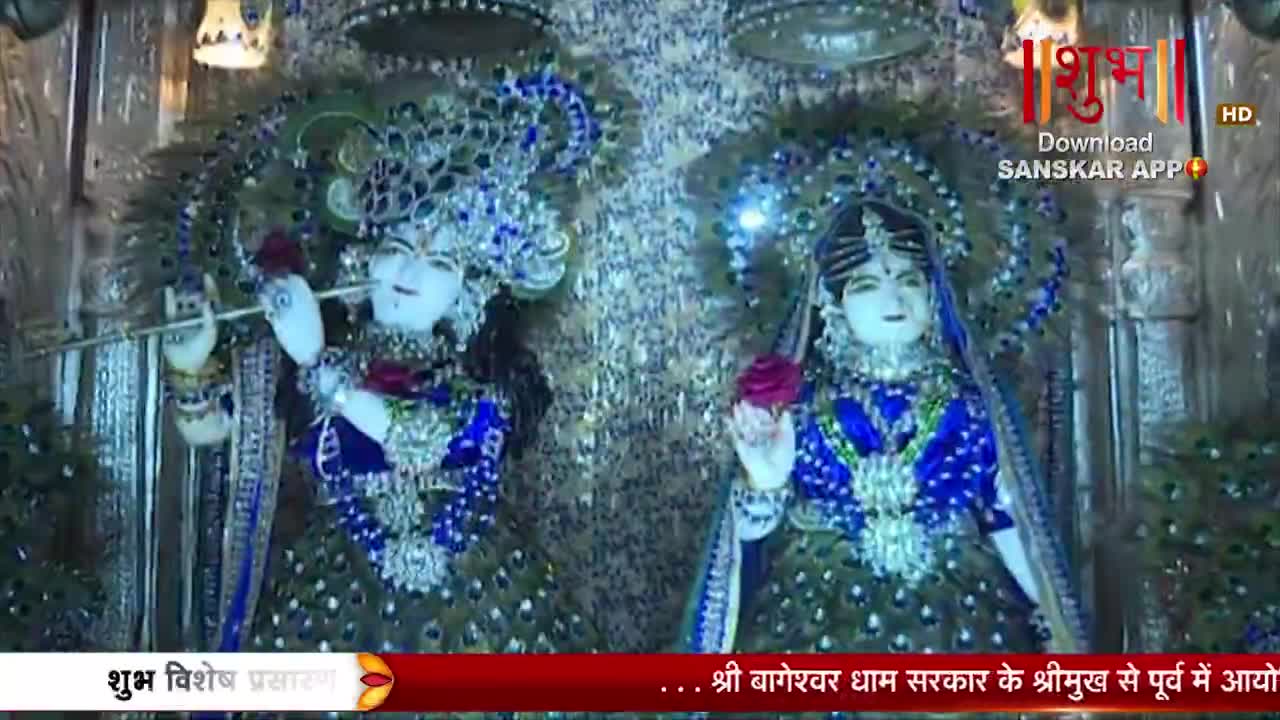 Two ornate statues of deities stand adorned in vibrant blue and green peacock feather-like garments. The one on the left holds a flute to its lips, while the one on the right extends a hand holding a rose.