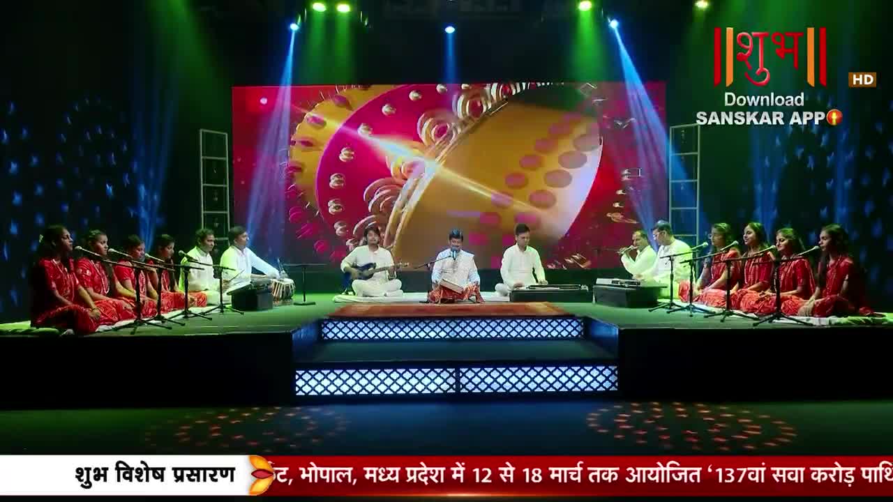A group of musicians performs on a stage bathed in colorful lights, with a large screen displaying abstract visuals behind them. A man in the center reads from a book while others play instruments, and a chorus of women in red saris sings.